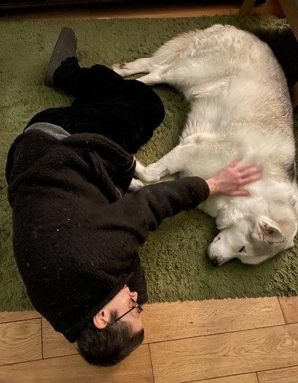 Zoose, our beautiful white husky (they all are), lying on a green rug, with my wife Cleodhna also lying on the rug, reaching out to him, in a yin-yang-style arrangement. They are both for the moment at peace.